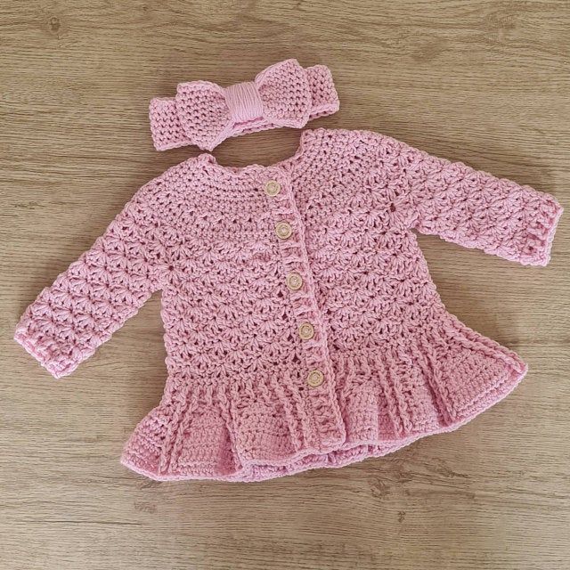 a pink crocheted cardigan with a bow on the front and bottom, sitting on a wooden surface