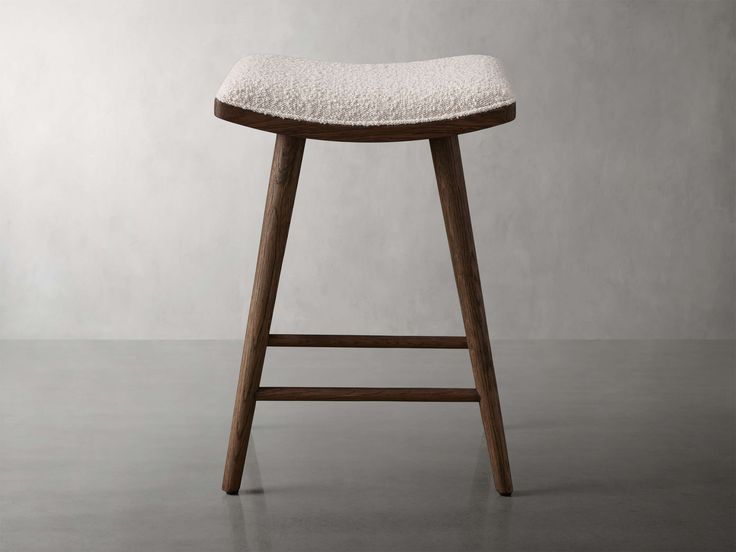 a wooden stool with a white upholstered seat and back rest, in front of a gray wall