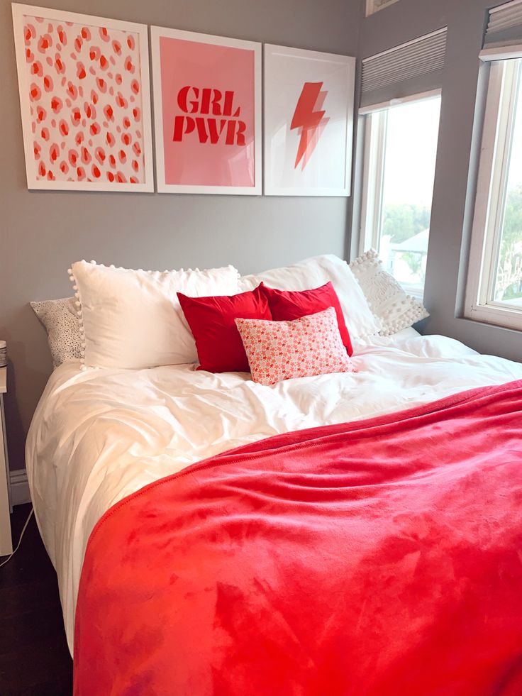 a bed with red and white pillows in a bedroom next to a window, framed pictures on the wall