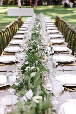 the long table is set with white plates and place settings for an outdoor dinner party