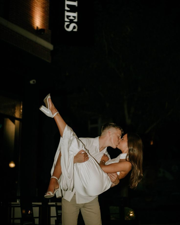 a man and woman are kissing in front of a building at night with their arms around each other