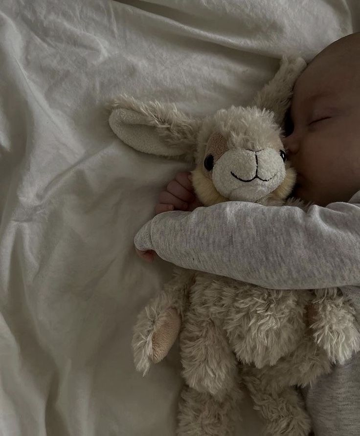 a baby sleeping next to a stuffed animal on a white sheet covered bed with it's head tucked under the pillow