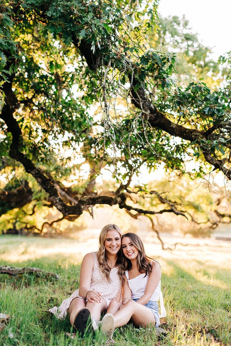 two women sitting in the grass under a tree with their arms around each other and smiling at the camera