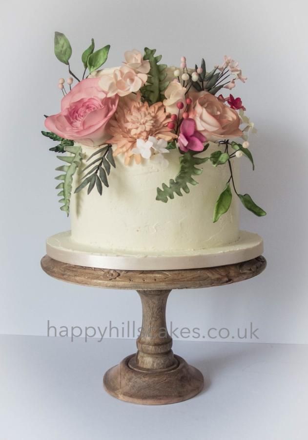 a white cake with pink flowers and greenery on top is sitting on a wooden stand