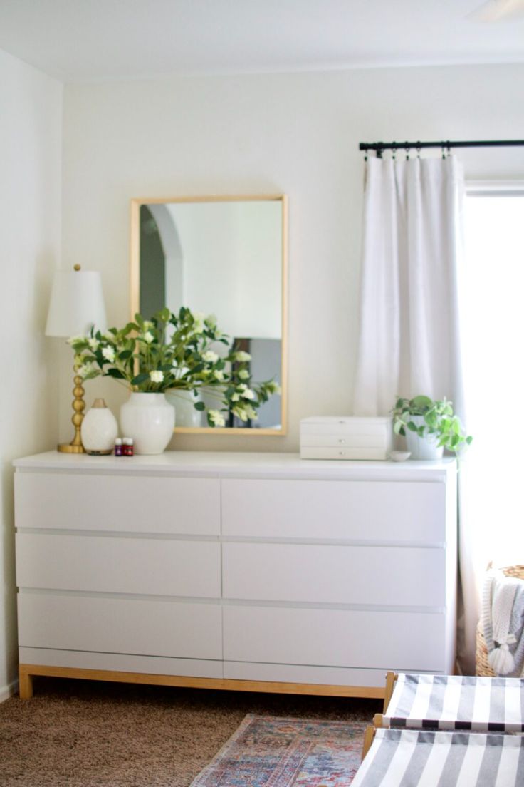 a white dresser sitting next to a window filled with plants and potted greenery