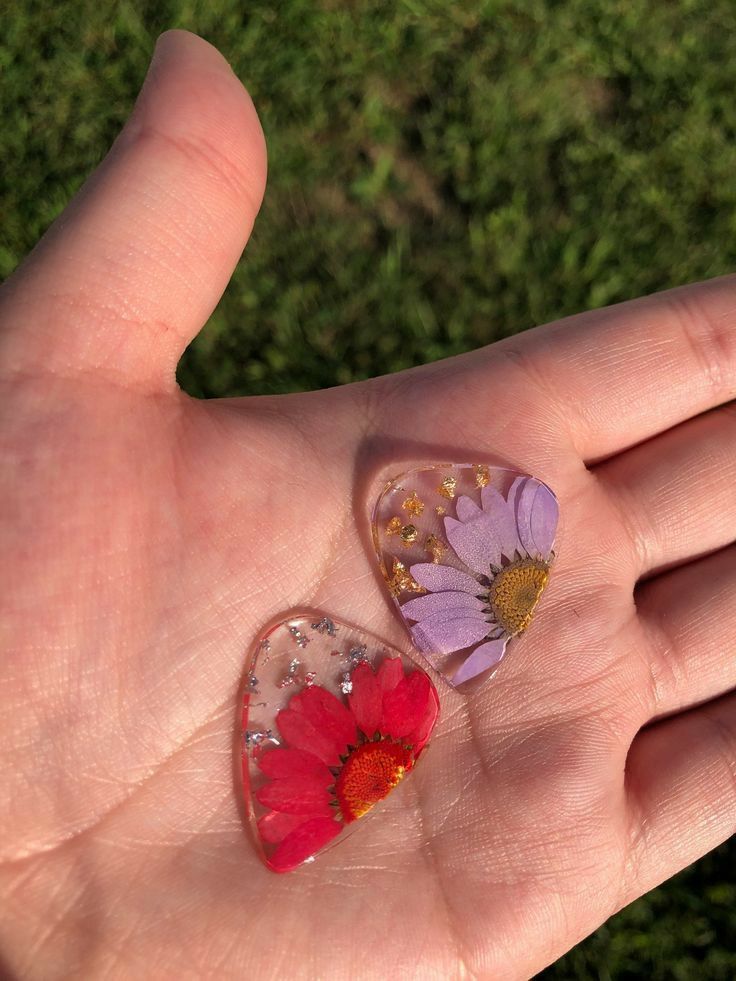 a person's hand holding two small pieces of glass with flowers on them and grass in the background