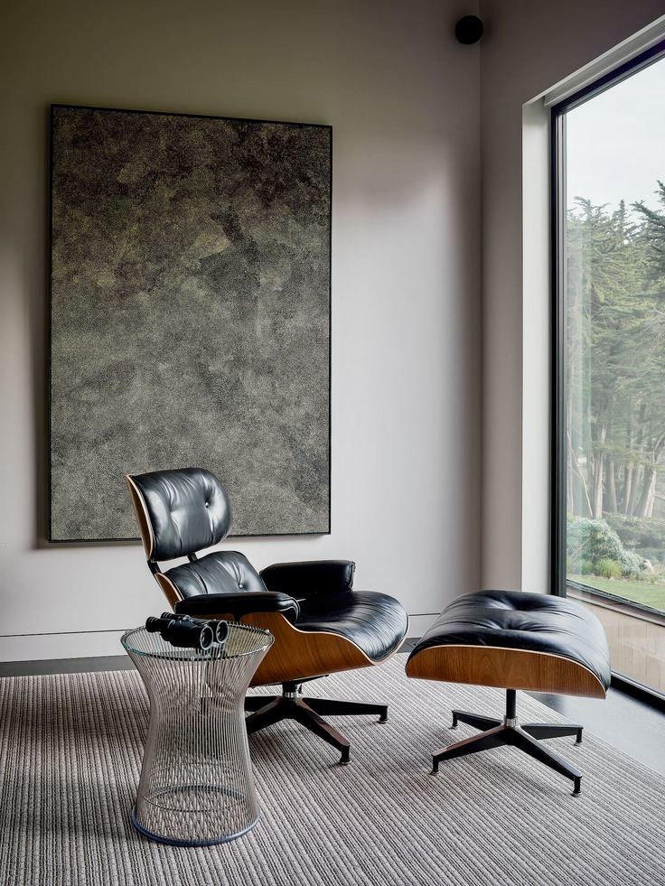 an eames chair and ottoman in front of a large window with a painting on the wall