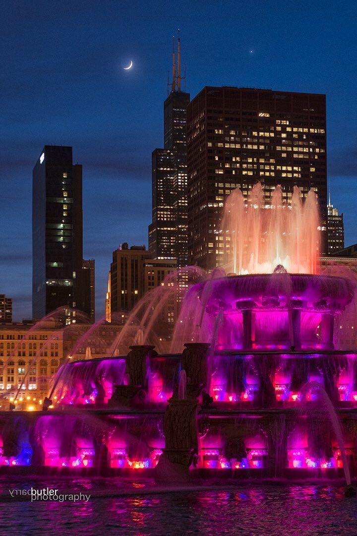 the water fountain is lit up with purple lights in front of the city's skyscrapers