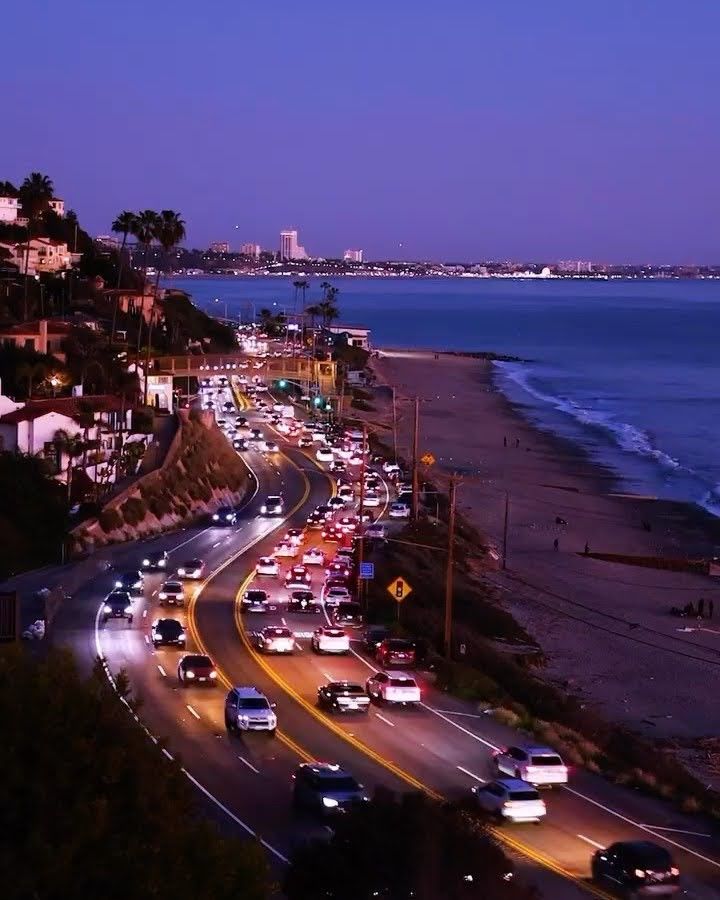 cars are driving down the road by the beach at night time with city lights in the background