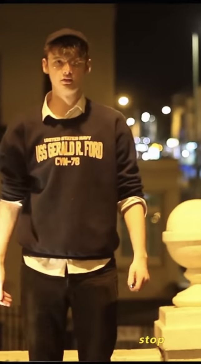 a young man standing in front of a white pillar wearing a black sweater and cap