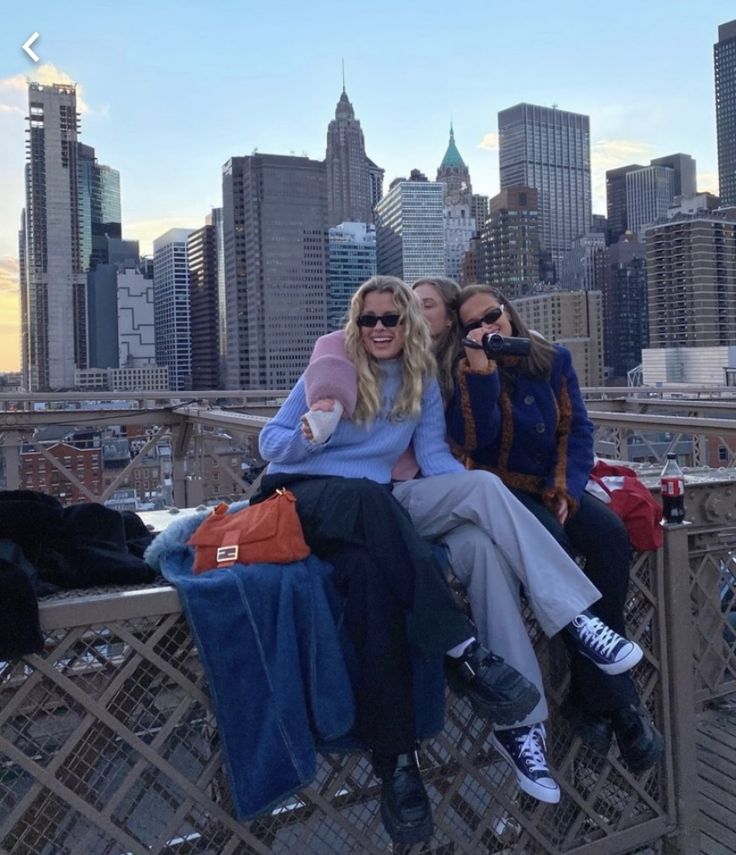 two women sitting on the edge of a bridge in front of a cityscape