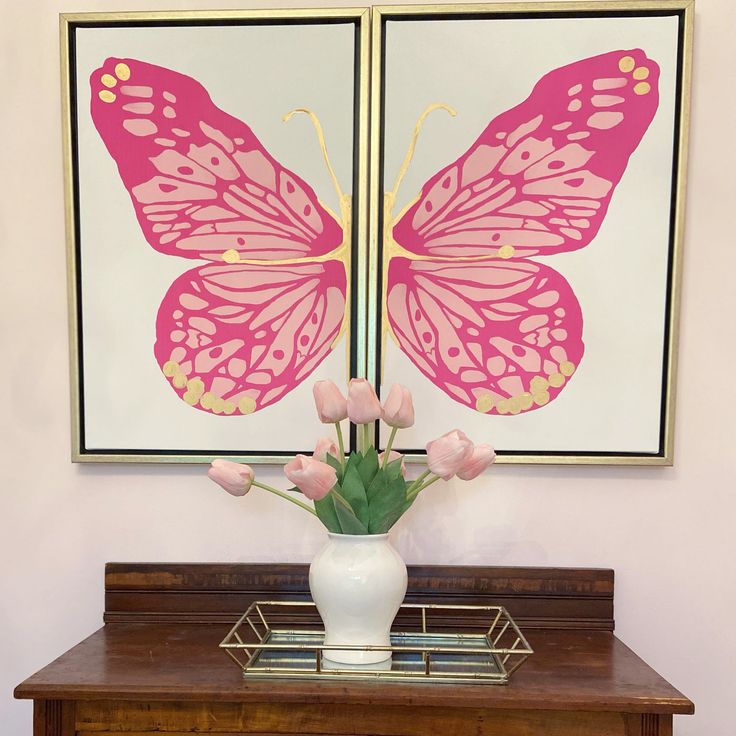 a white vase with pink flowers on top of a wooden table next to a painting