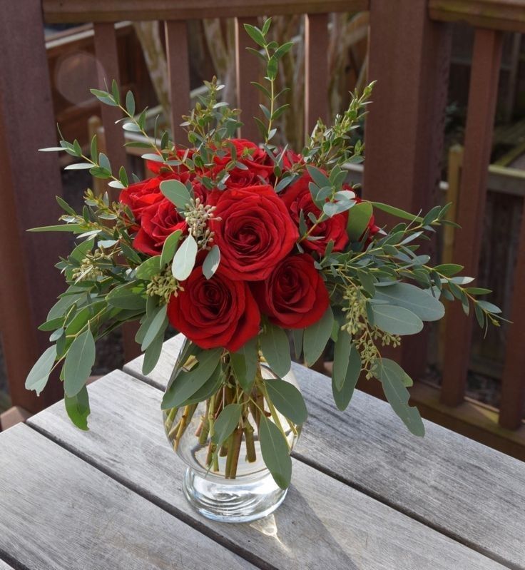 a vase filled with red roses and greenery on top of a wooden table next to a fence