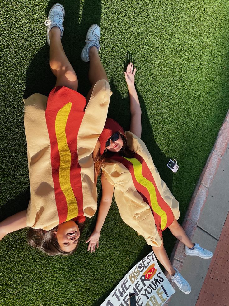 two women dressed as hot dogs laying on the grass with their hands in the air