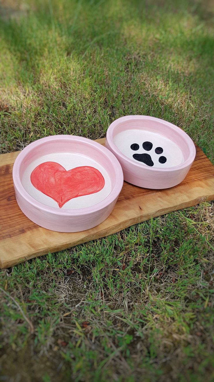 two bowls with hearts painted on them sitting on a wooden board in the middle of grass