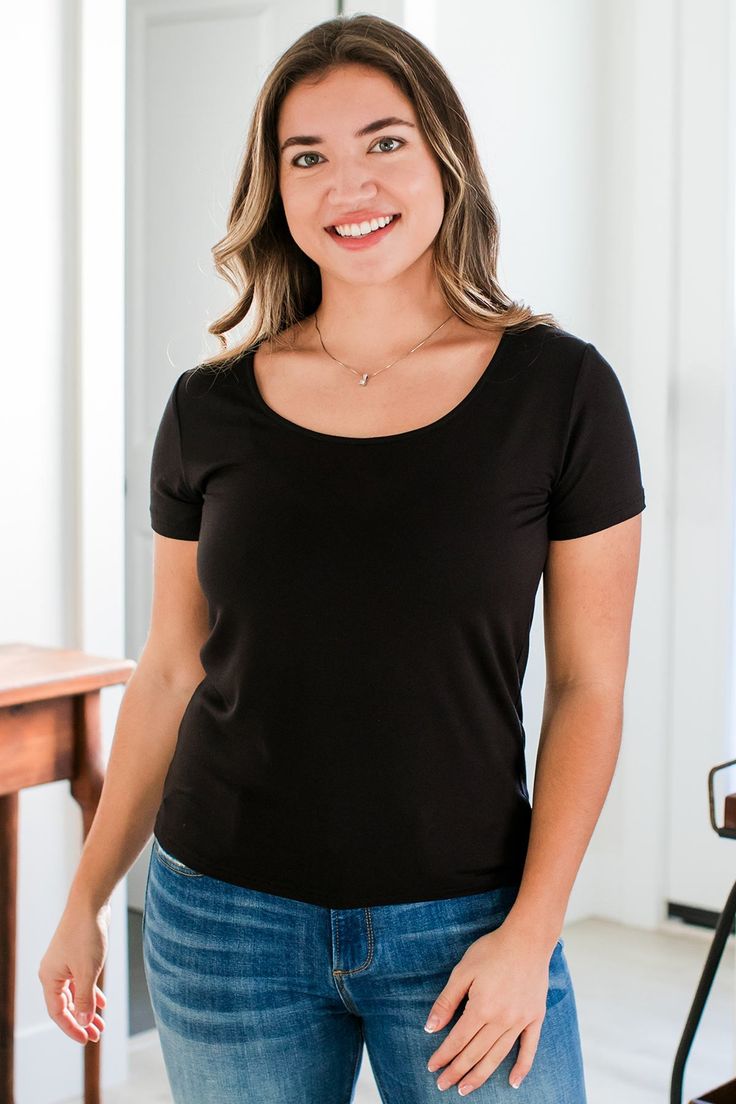 A woman standing with one hand lightly resting on the front of her thigh Everyday Stretch T-shirt With Scoop Neck, Fitted Scoop Neck Short Sleeve Top, Fitted Short Sleeve Scoop Neck Top, Basic Fitted Short Sleeve Top With Scoop Neck, Scoop Neck Fitted T-shirt For Everyday, Fitted Scoop Neck T-shirt For Everyday, Classic Fitted Scoop Neck T-shirt, Stretch Scoop Neck T-shirt For Everyday, Black Scoop Neck T-shirt For Everyday