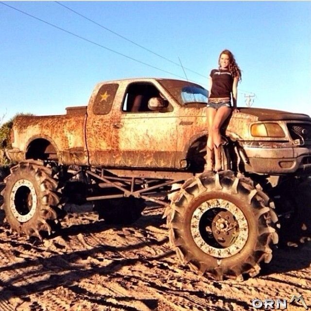 a woman sitting on the back of an old truck with large tires in front of her