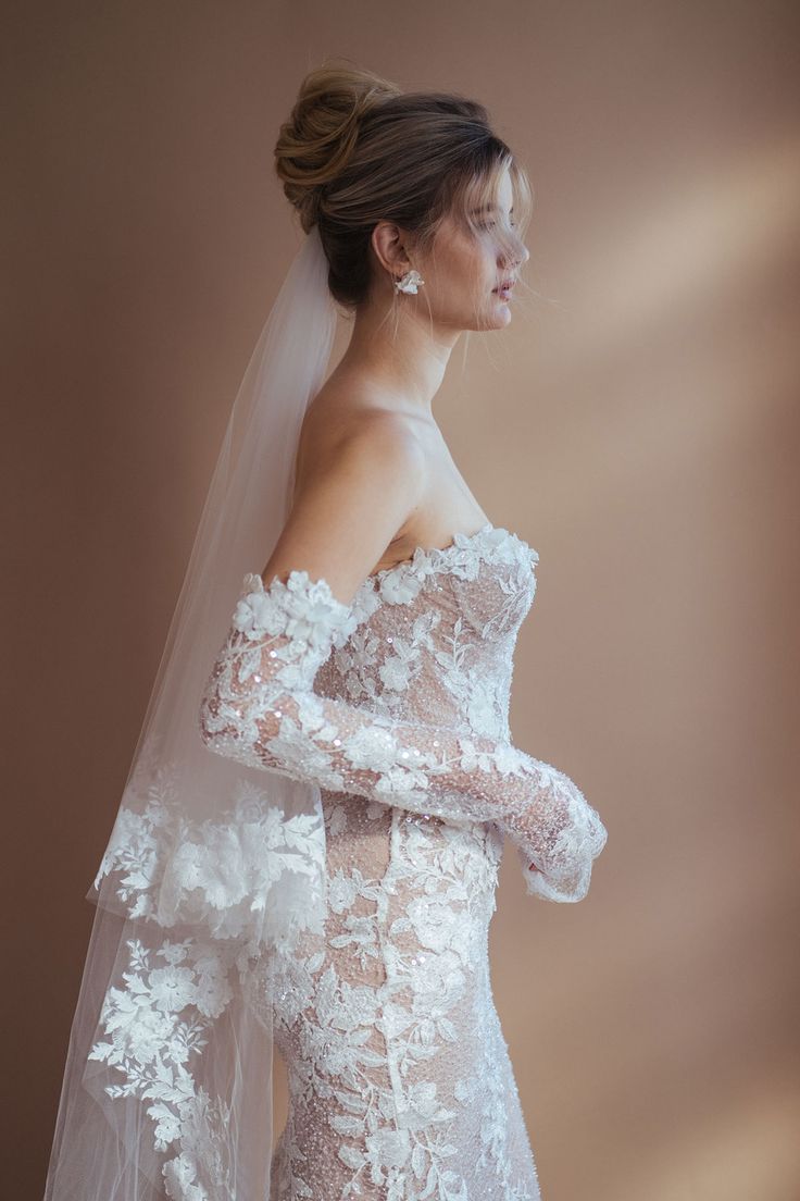 a woman wearing a wedding dress with long sleeves and flowers on the shoulder, standing in front of a brown background