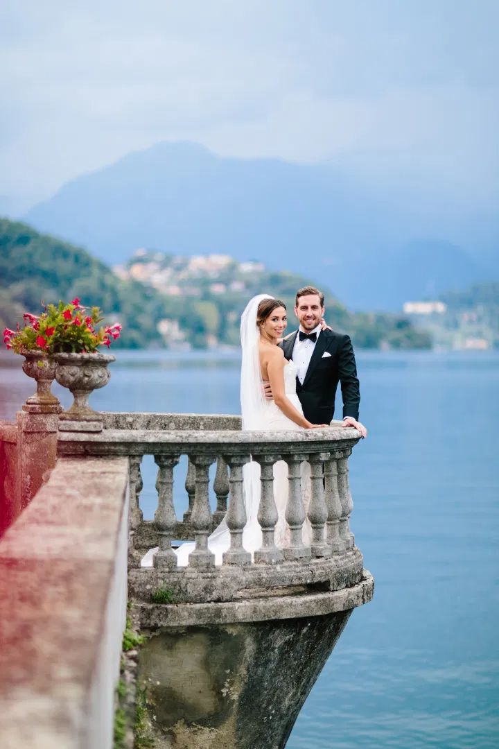 a bride and groom standing on the edge of a balcony next to a body of water