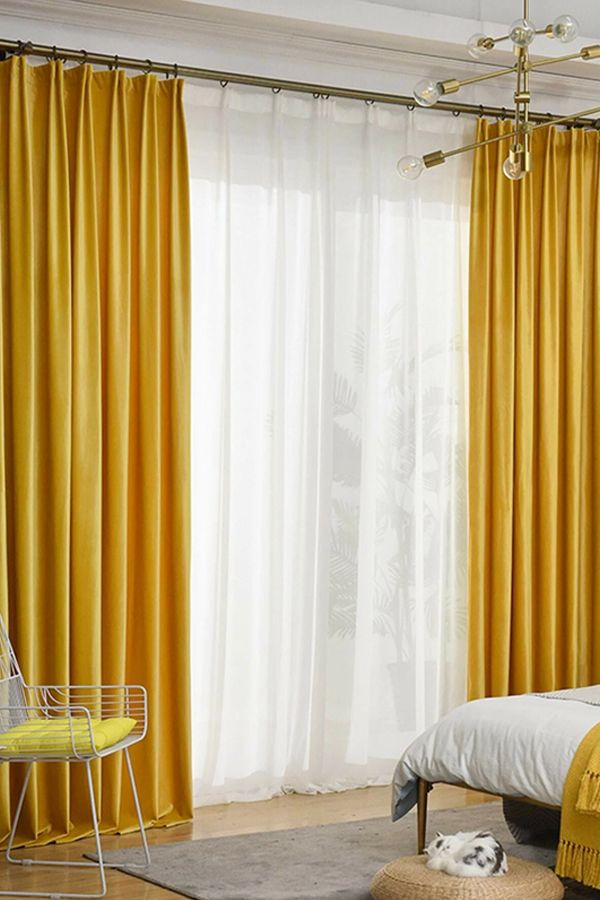 a bedroom with yellow curtains and a white bed in front of a window that has a dog laying on the floor next to it