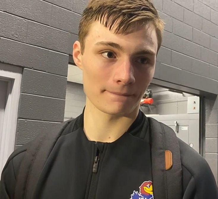 a young man standing in front of a gray brick wall wearing a black jacket with an eagle patch on it