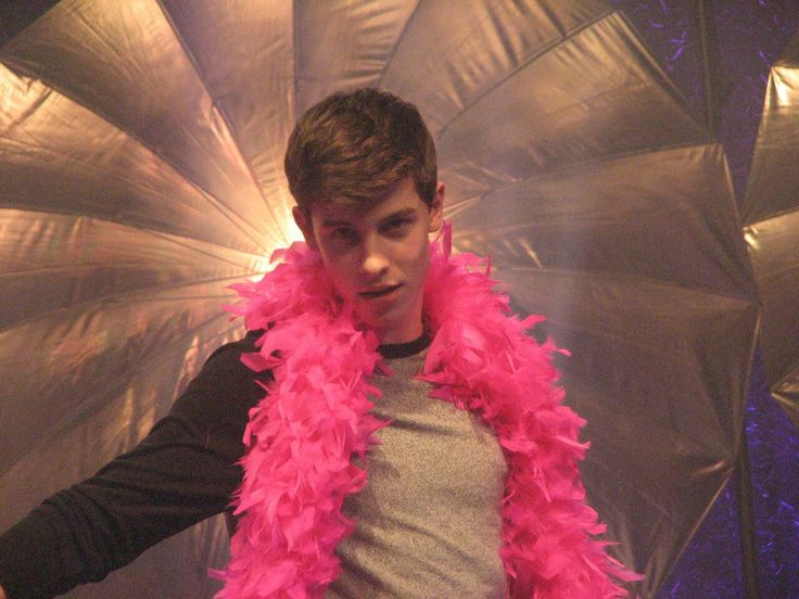 a young man wearing a pink feather boa around his neck and shoulders, standing in front of a silver background