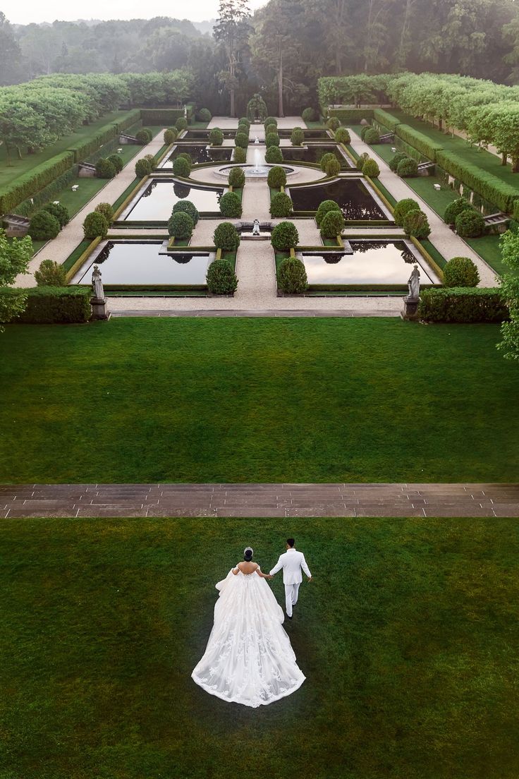 the bride and groom are walking in front of an elaborate formal garden with water features