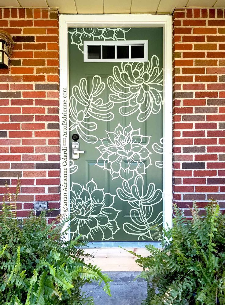 a green door with white flowers painted on the glass and brick wall next to plants