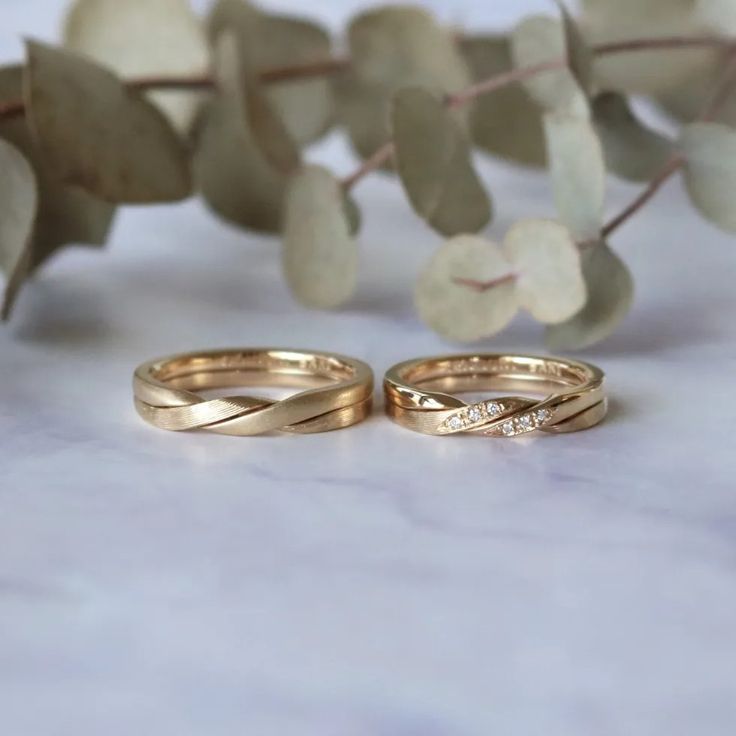 two gold wedding rings sitting next to each other on a white marble surface with greenery in the background