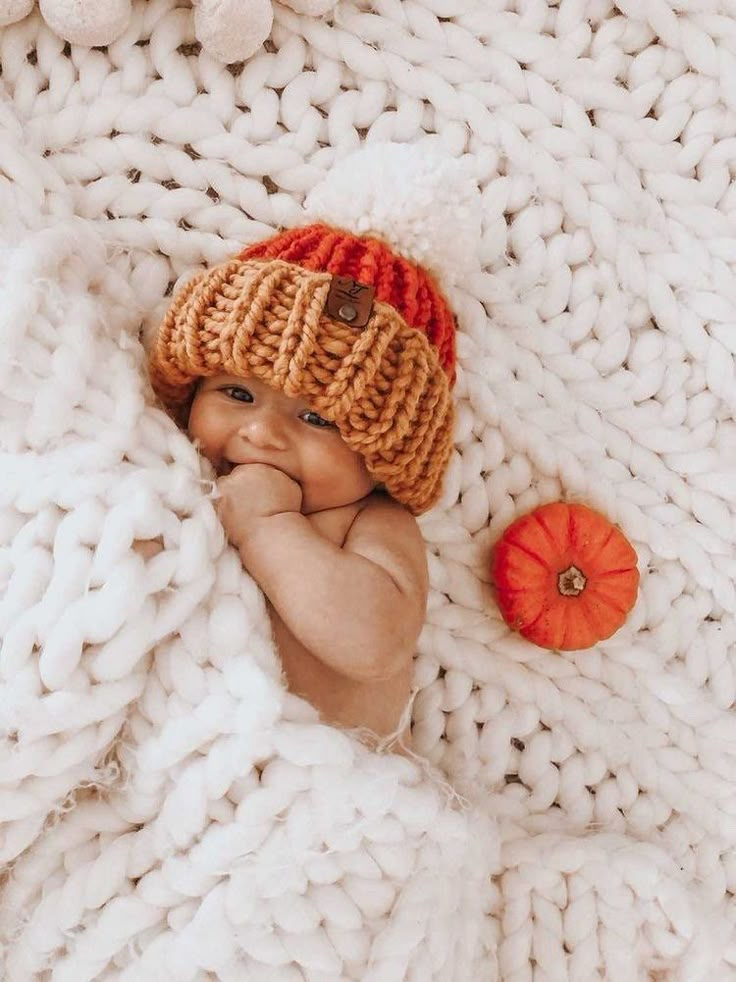 a baby wearing a knitted hat laying on top of a blanket next to an orange pumpkin