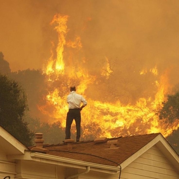 a man standing on top of a roof in front of a fire raging through the sky