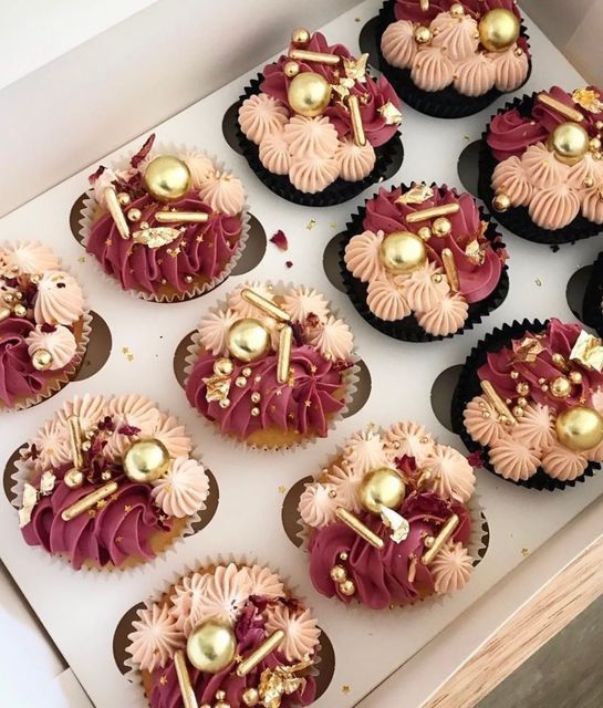 twelve cupcakes with pink frosting and gold decorations in a white box on top of a wooden table