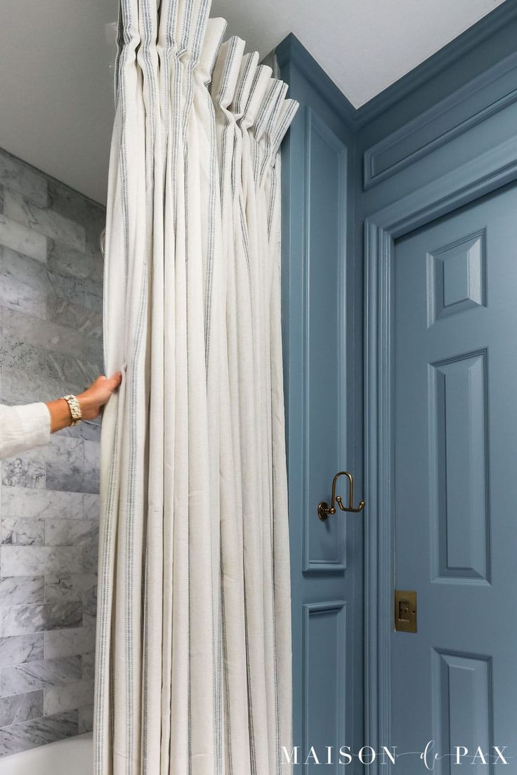 a woman is opening the curtains in front of a blue door and a white bathtub