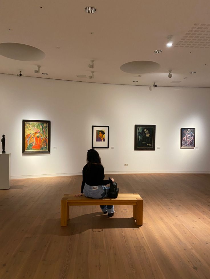 a woman sitting on a bench in an art gallery looking at paintings and artwork hanging on the walls