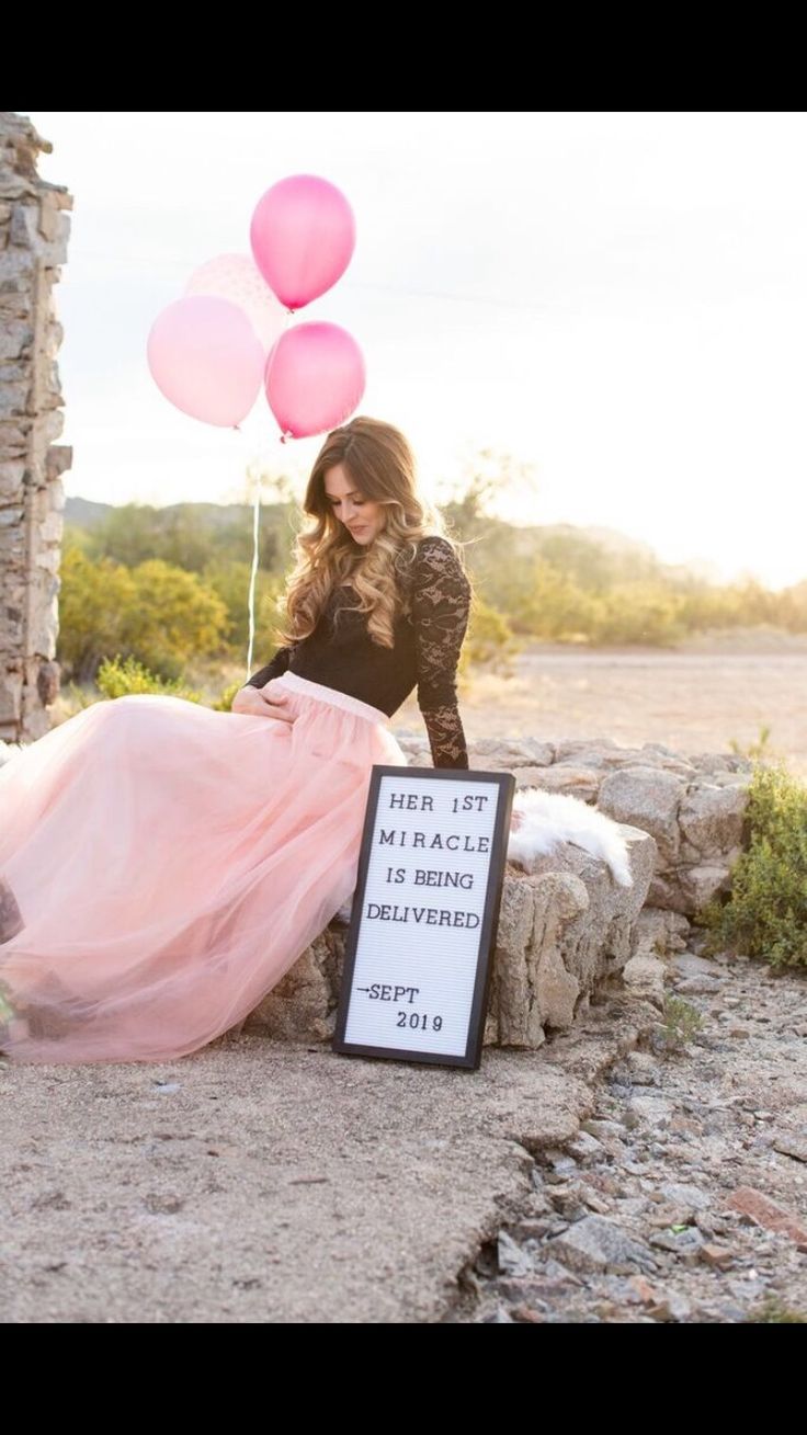 a woman in a long pink skirt sitting next to a sign with balloons on it