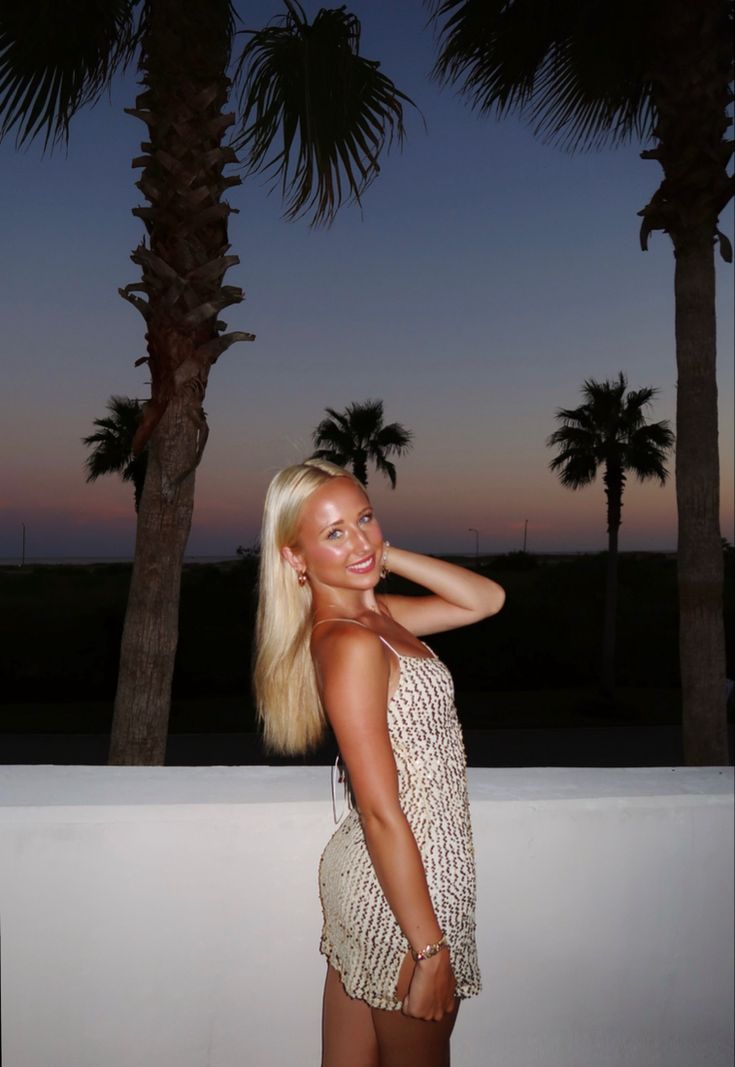 a woman standing next to palm trees at night with her hand on her head and smiling