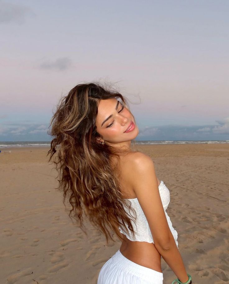 a beautiful young woman standing on top of a sandy beach next to the ocean with her hair blowing in the wind