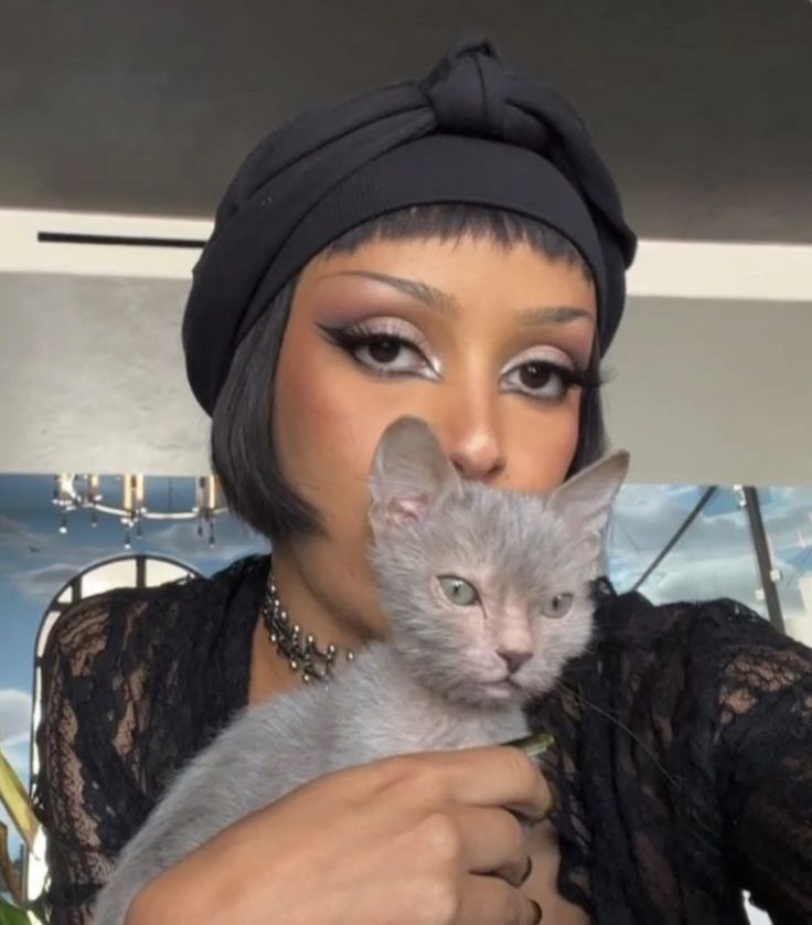 a woman holding a gray cat in her hands while wearing a turban hat