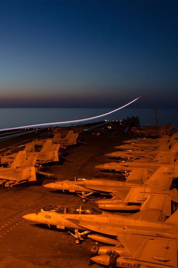 an air port filled with lots of fighter jets at night, and one plane is flying in the sky