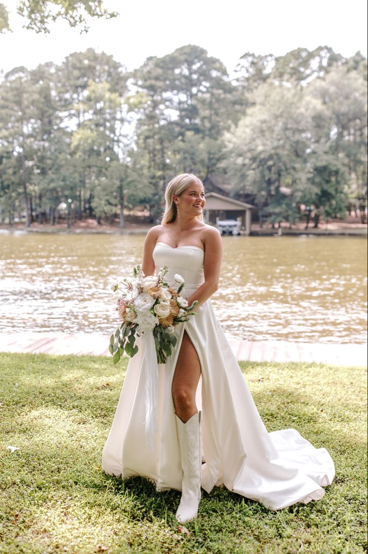 a woman standing in front of a body of water wearing a white dress and cowboy boots