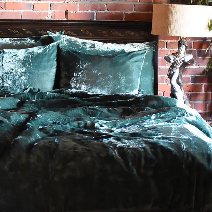 a bed with green comforter and pillows in front of a brick wall next to a lamp