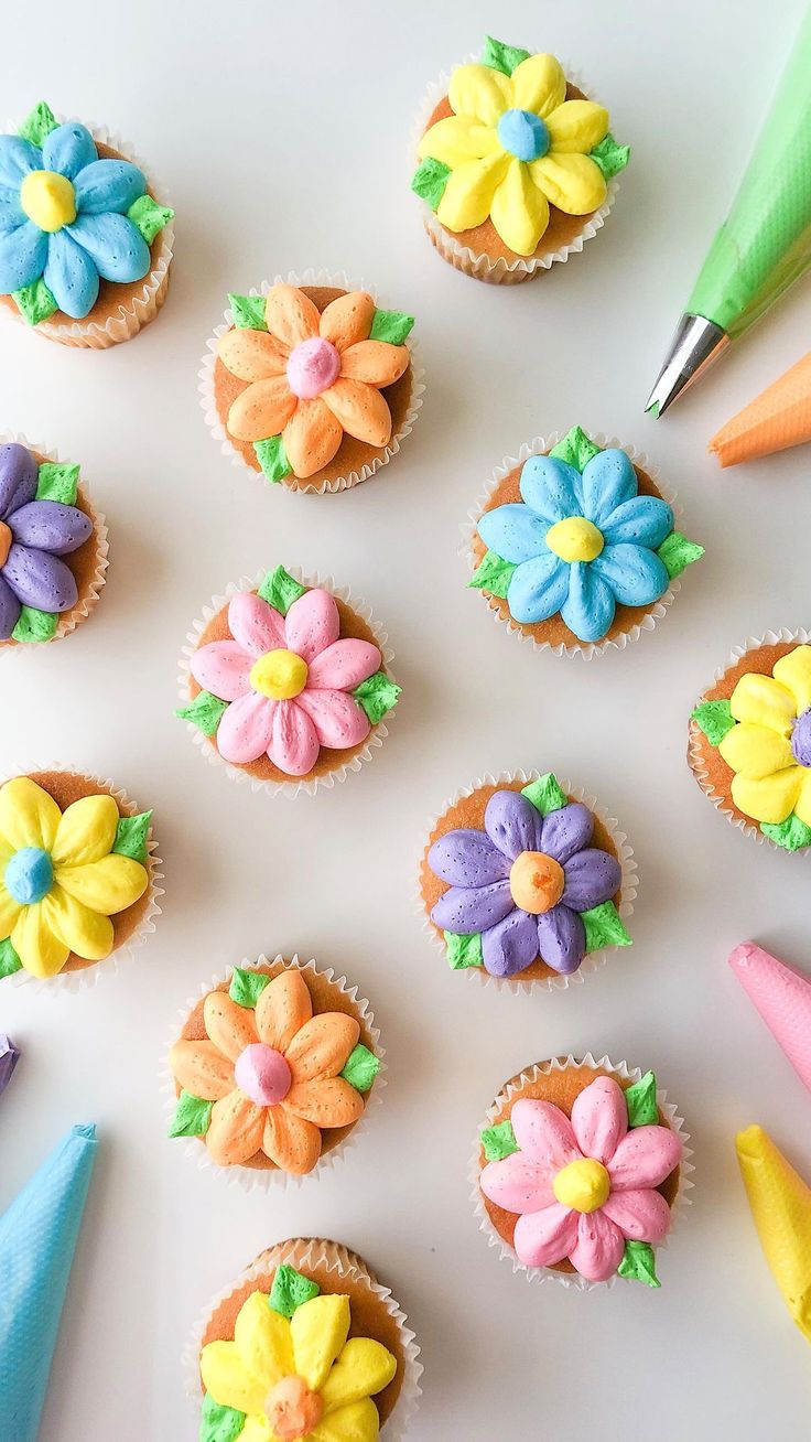 cupcakes decorated with icing and flowers are arranged on a white surface next to colored crayons