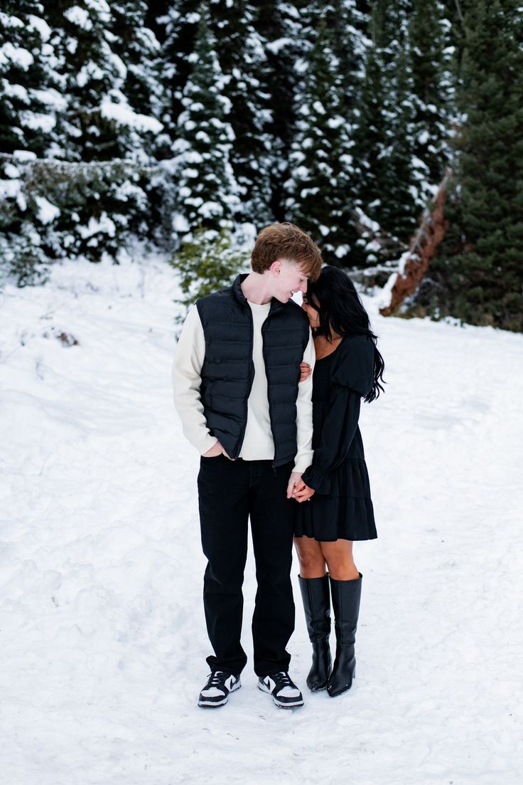 Man and woman standing close together in the snow. The man is smiling down at the woman and the woman is kissing his shoulder. Outfits For Engagement Pictures Winter, Winter Proposal Outfit, Winter Engagement Outfits, Winter Engagement Photos Outfits, Winter Proposal, Winter Engagement Pictures, Engagement Photos Outfits, Engagement Picture Outfits, Forest Engagement