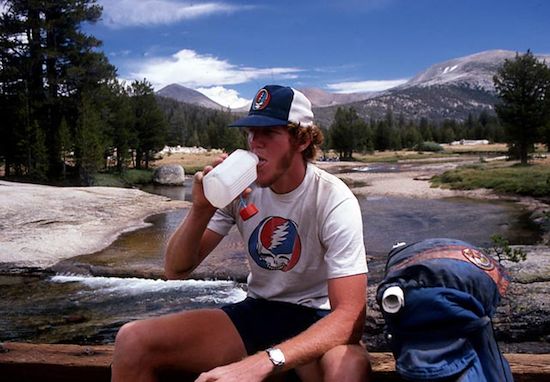 a man sitting next to a river drinking from a paper cup while wearing a hat