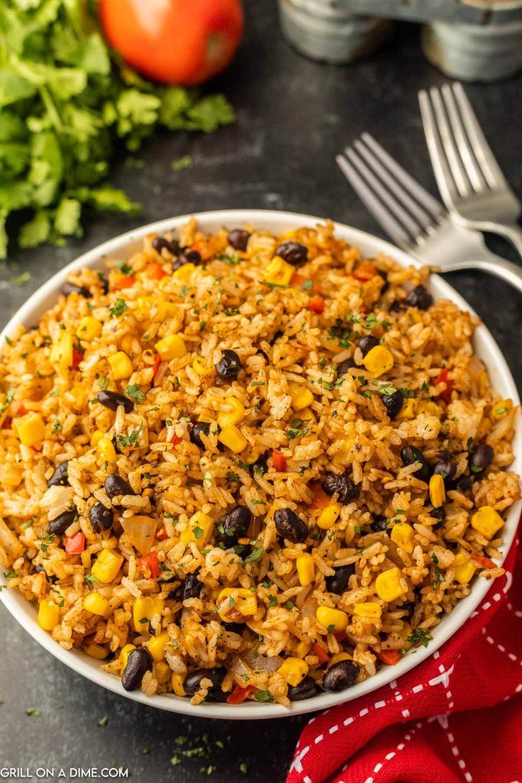 mexican rice and beans in a white bowl on a black surface with silverware, tomatoes, and lettuce