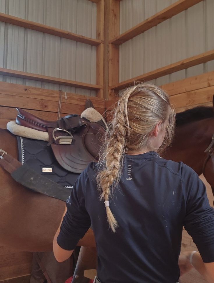 a woman standing next to a brown horse in a stable with saddles on it's back