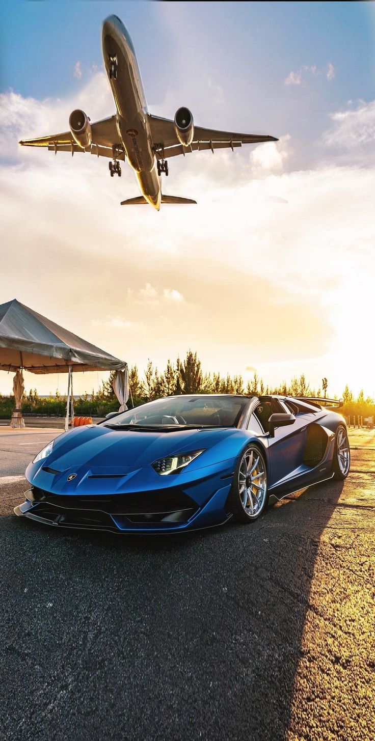 a large jetliner flying over a blue sports car in front of a parked lamb