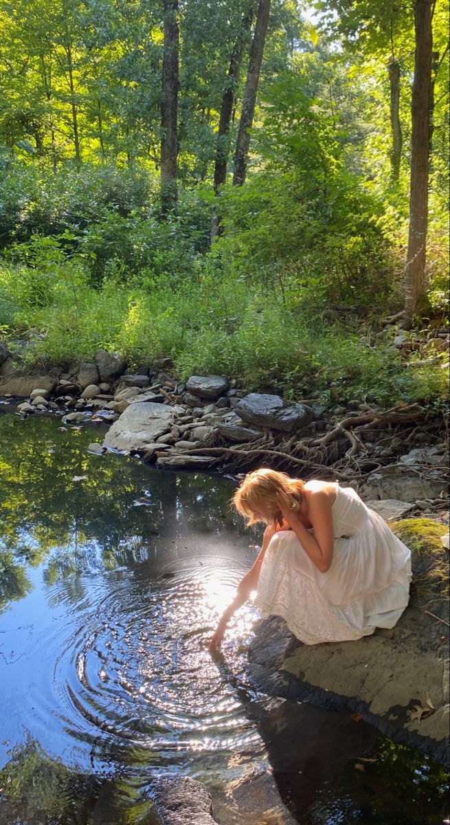 a woman in a white dress is sitting on rocks by the water and looking at her reflection