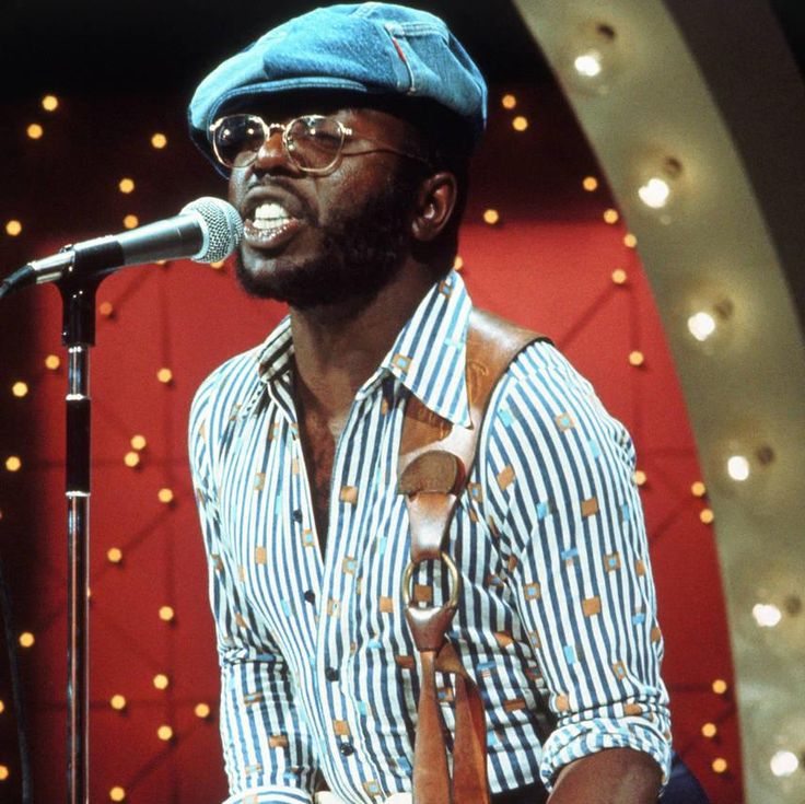 a man standing in front of a microphone with a hat on his head and holding a guitar