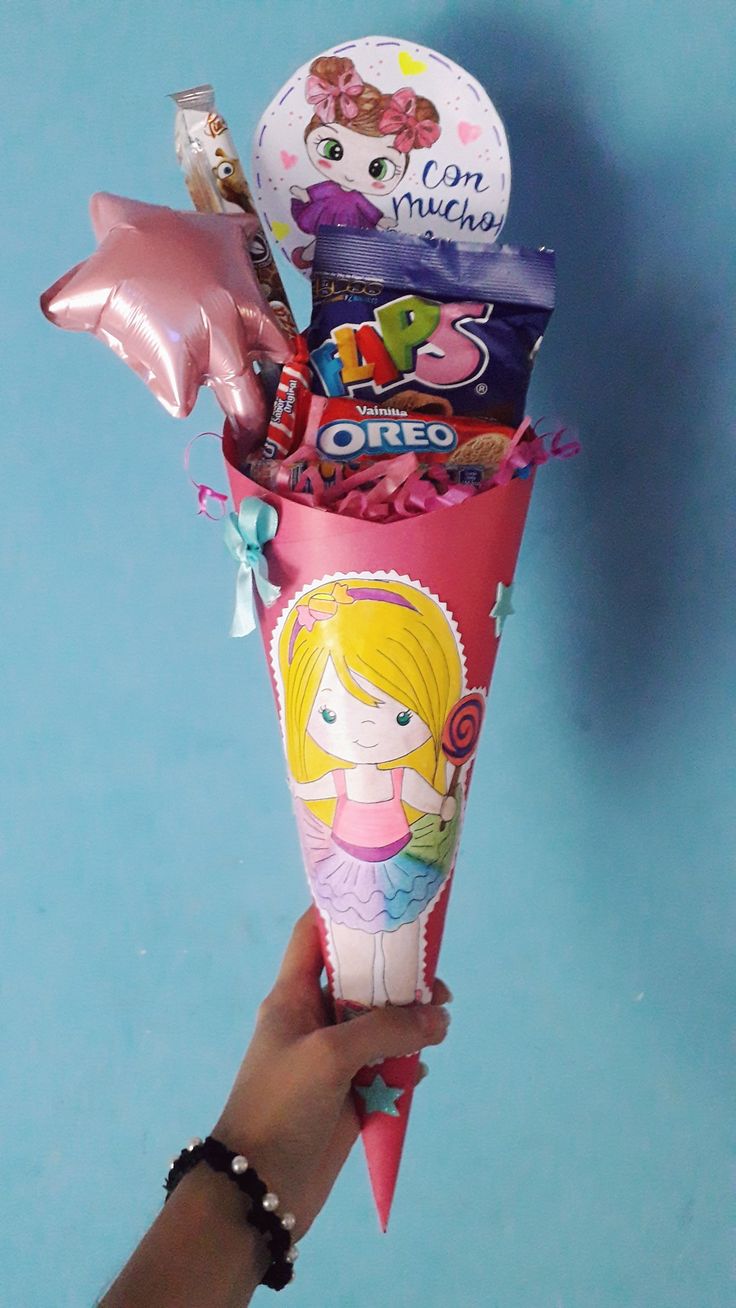 a hand holding a pink cone filled with lots of candy and balloons on top of a blue surface